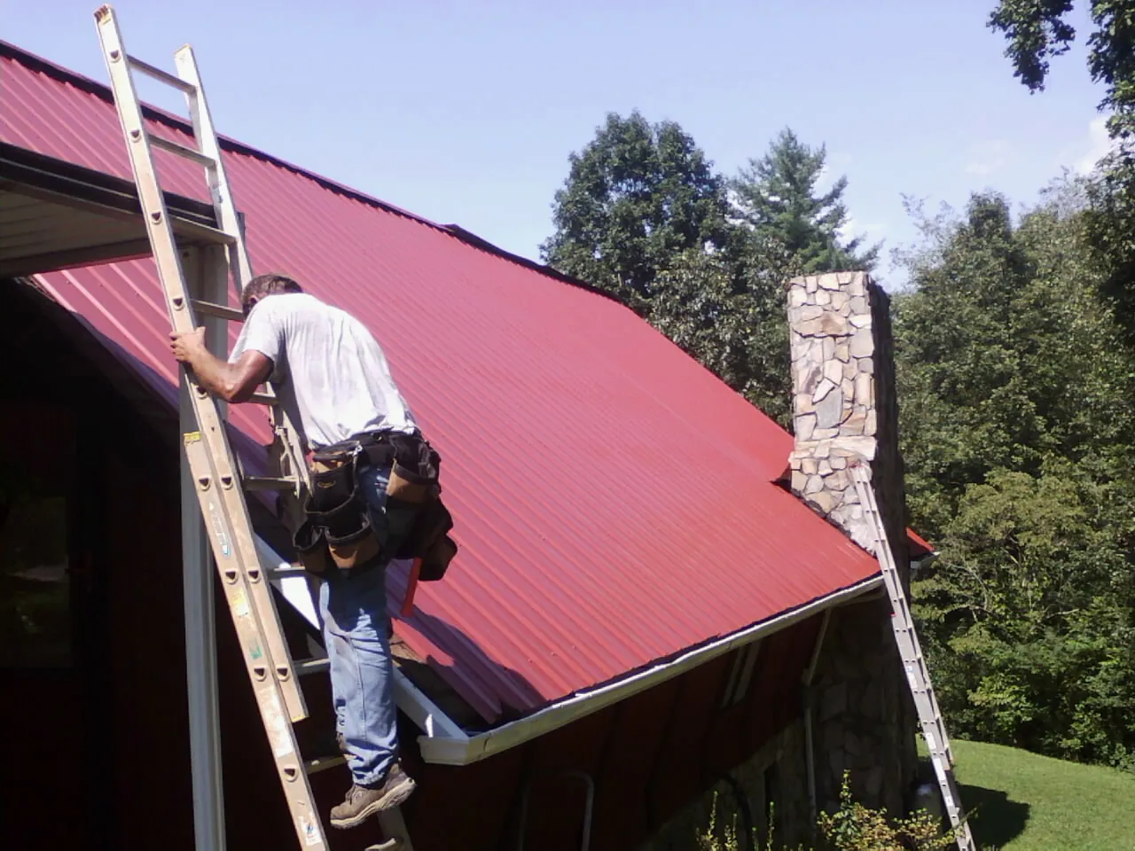Roofer on a ladder beside a red metal roof and stone chimney — Soffit & Fascia Repair by Roofing Brothers in Maquon